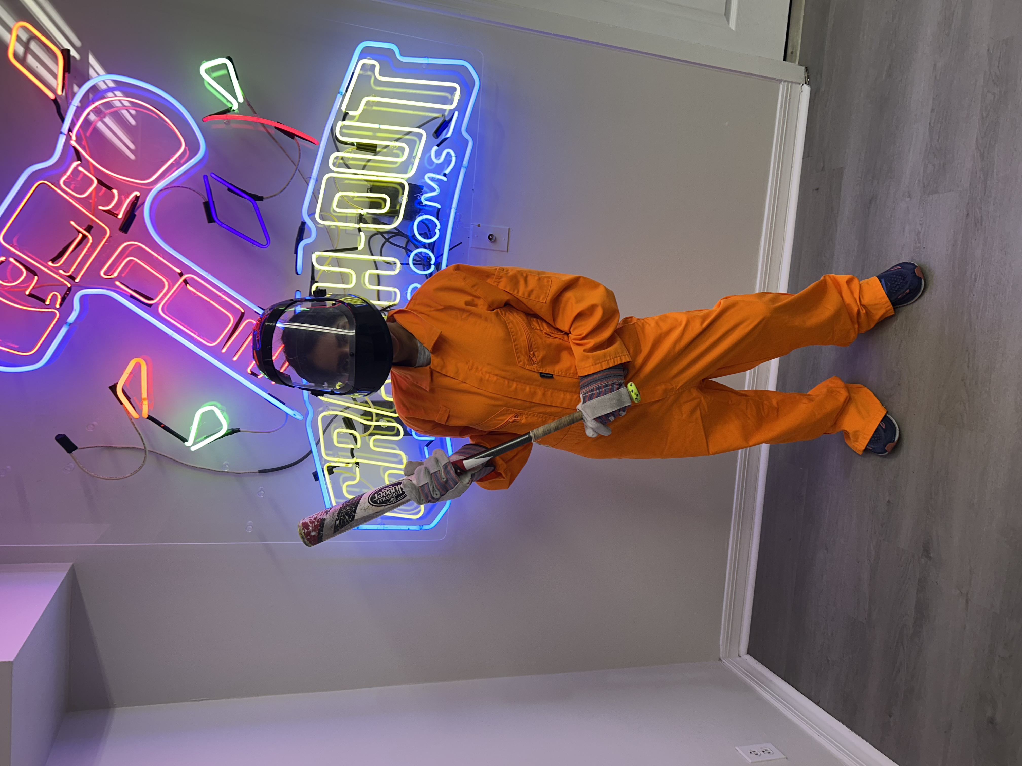 Crash Out customer in orange jumpsuit posing with bat in front of vibrant Crash Out Rage Rooms neon sign