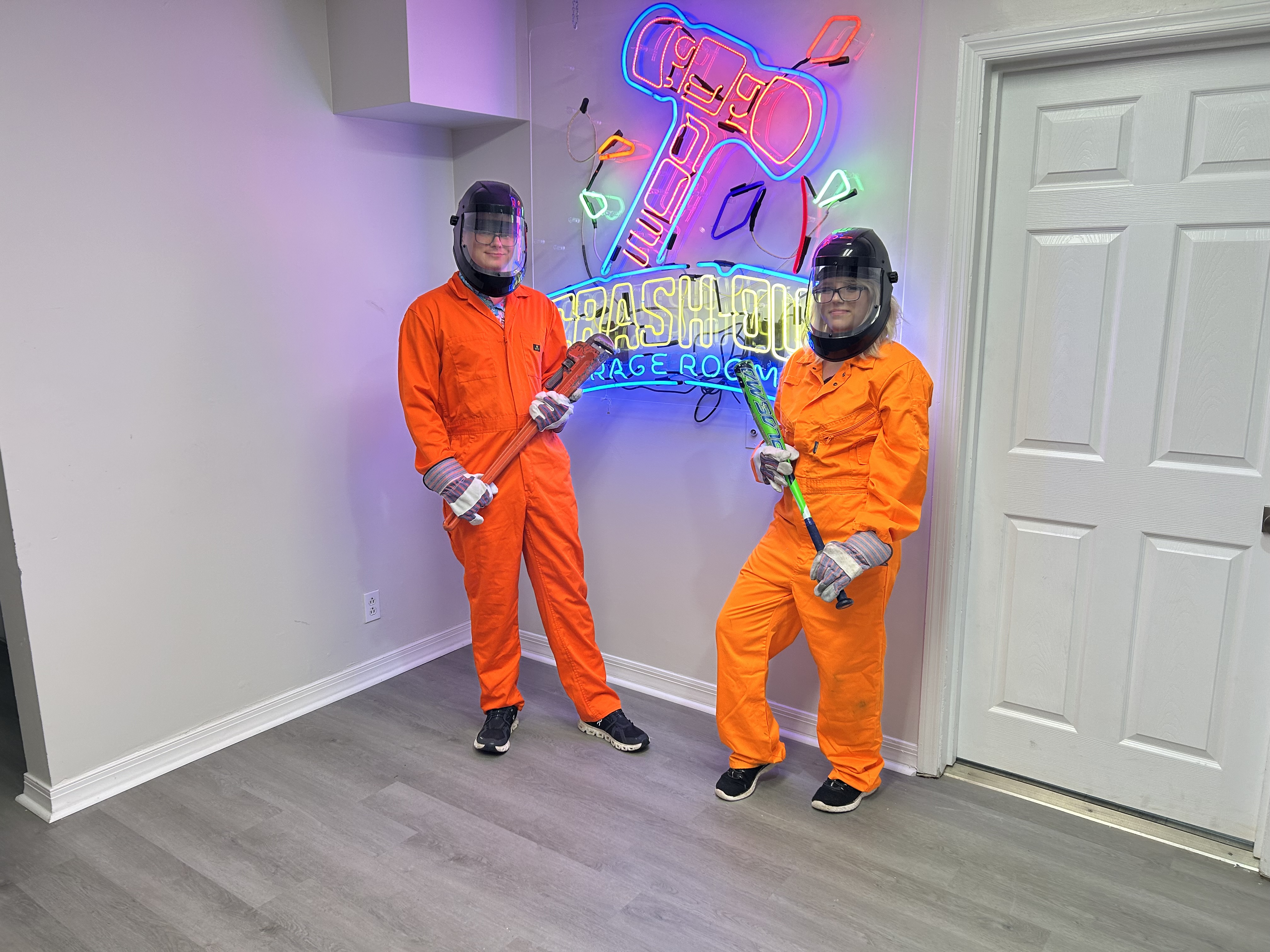 Large group of Crash Out Rage Rooms guests in orange jumpsuits holding bats in front of branded neon sign